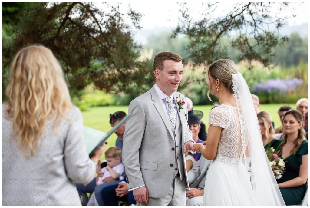 The bride it putting the ring on the grooms finger - smiling guests are watching in the background